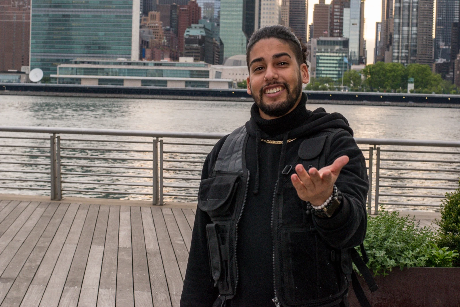 Young man reaching toward the camera on a waterfront deck with the skyline behind