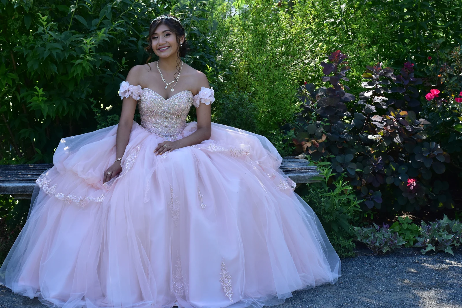 Quinceañera seated in her gown on a garden bench among summer greenery