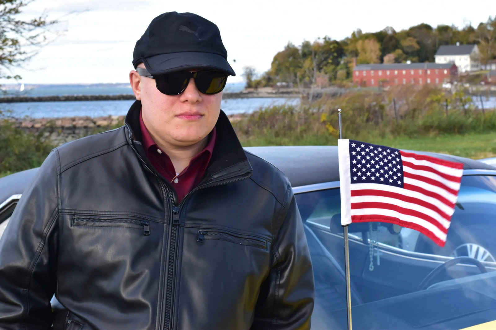 Man in a leather jacket and cap beside a small flag at the waterfront