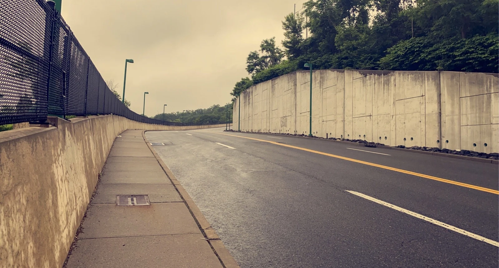 An empty road beside a concrete wall and fence on an overcast day