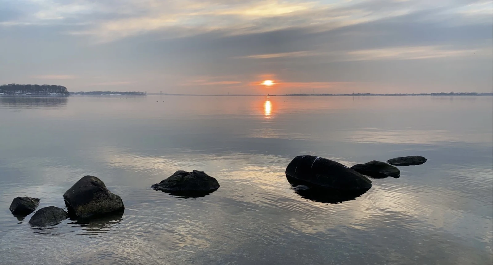 Sunrise over still water with dark rocks in the foreground
